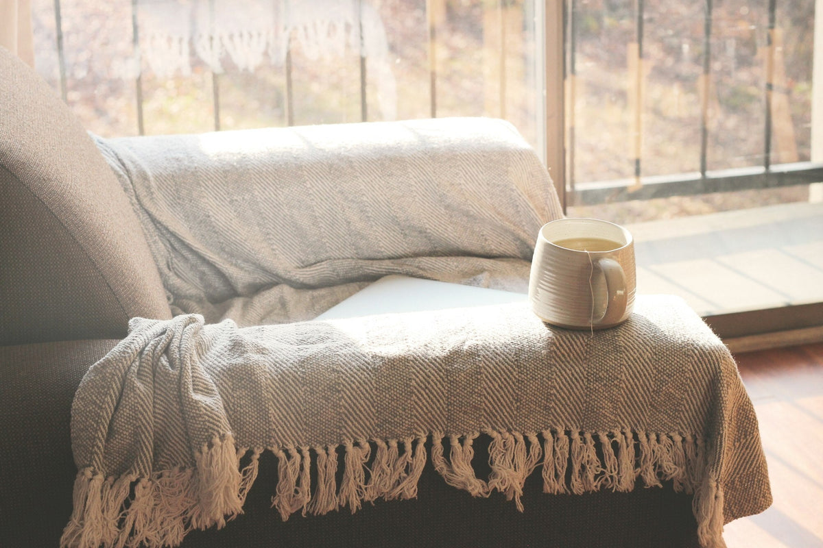 Tasse Tee auf einer Sofalehne mit heller Wolldecke am sonnigen Fenster
