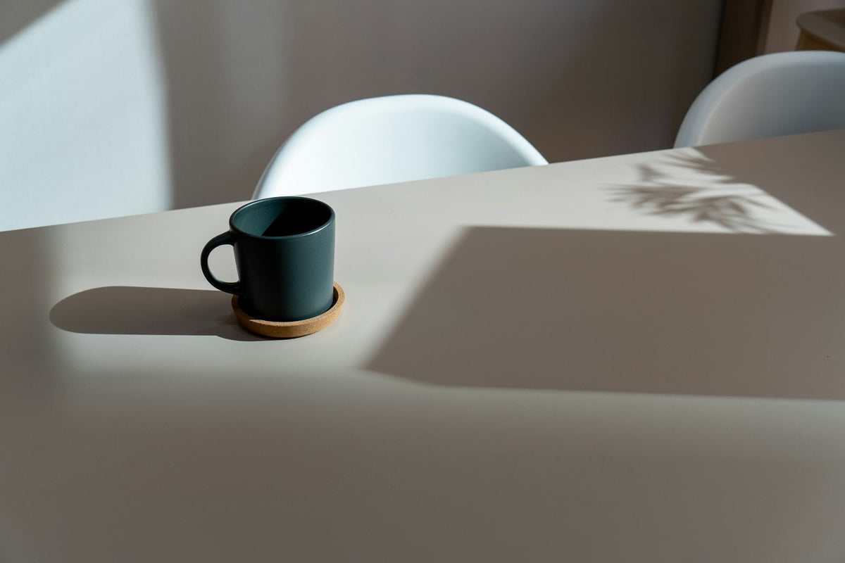 Becher auf Korkuntersetzer auf hellem Tisch im warmen Morgenlicht, daneben der Schatten einer Pflanze