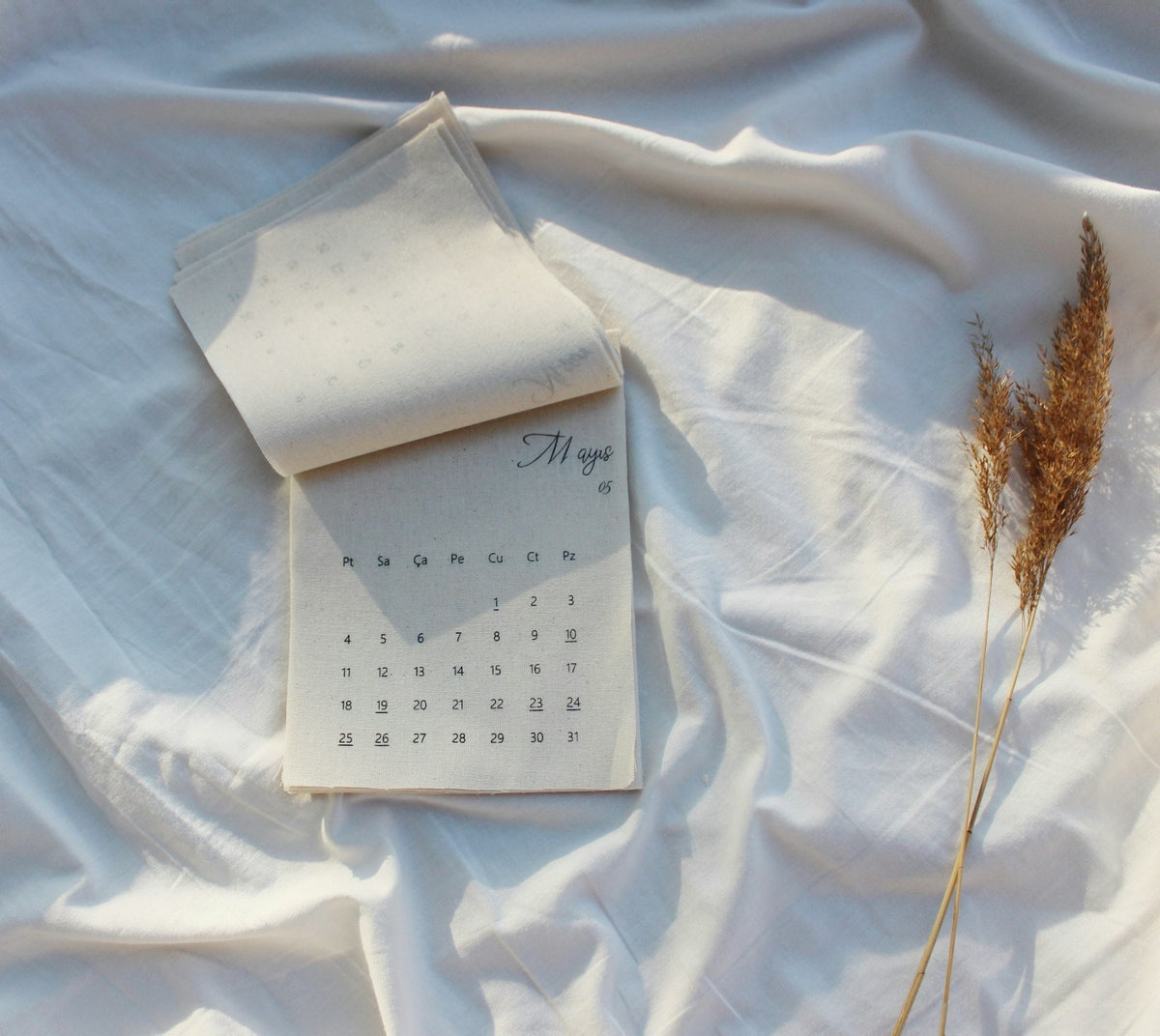 Aufgeschlagener Stoffkalender mit Monatsübersicht auf weißem Laken, daneben trockene Gräser